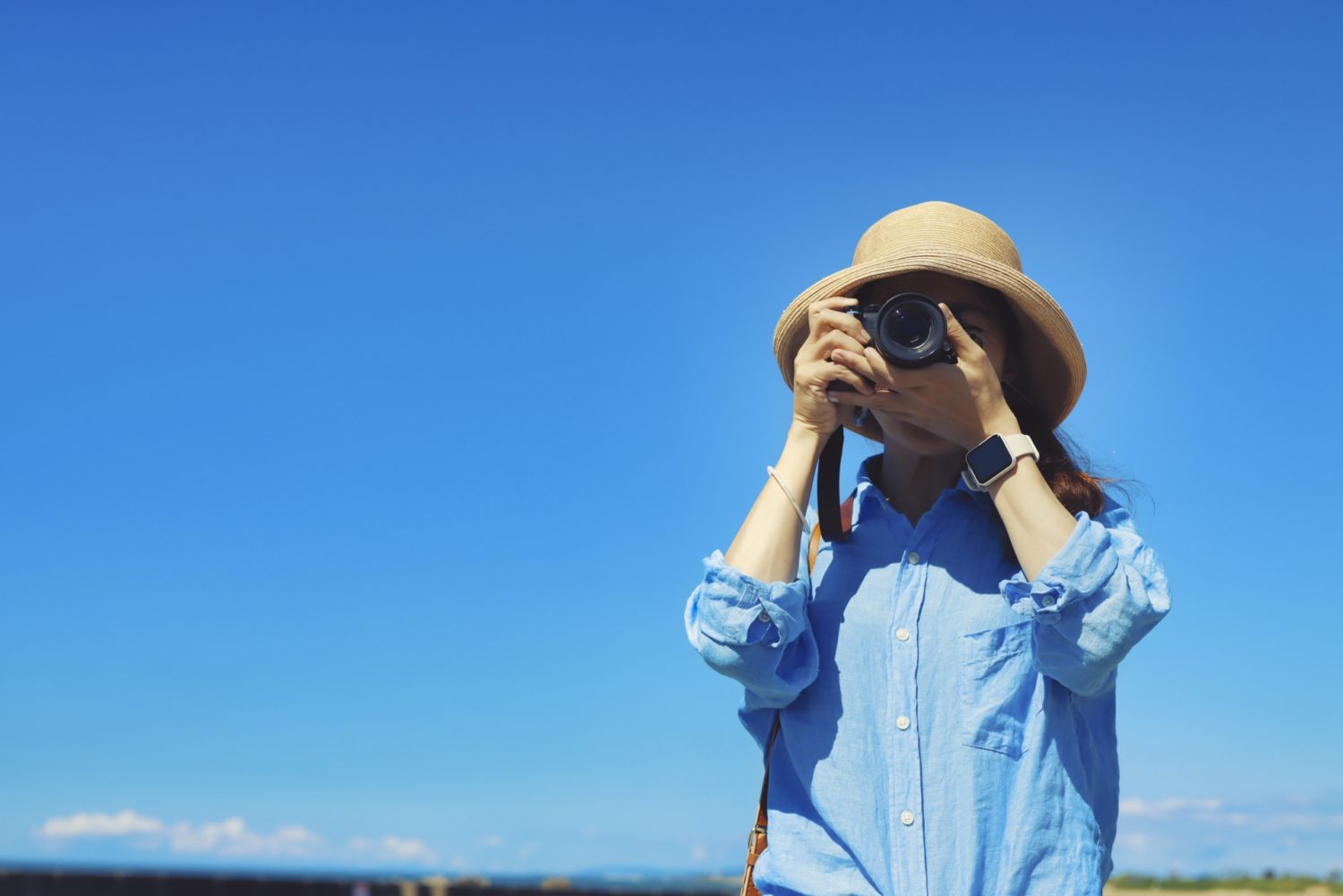 青空の下、女性がカメラで写真を撮影している様子。写真ACでの副業をイメージ