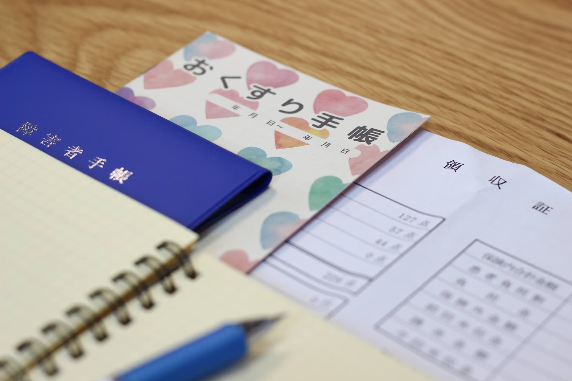 お薬手帳、障害者手帳、領収証の上に置かれたメモ帳とペン。発達障害の人が使える支援制度のイメージ。